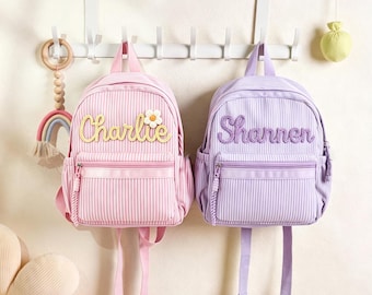 Può includere: Due zaini a righe, uno rosa e uno viola, con una tasca frontale con cerniera e una maniglia superiore. Lo zaino rosa mostra il nome Charlie in corsivo giallo con una margherita, mentre lo zaino viola riporta il nome Shannen in colore viola.