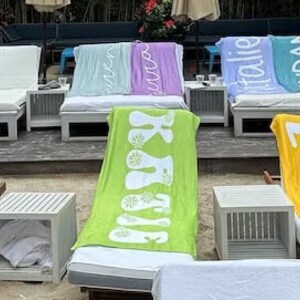 Pu&ograve; includere: Una fila di asciugamani da spiaggia colorati con diverse parole stampate su di essi, tra cui "Summer", "Basking", "Friday", "Italian" e "Brendan". Gli asciugamani sono disposti su sedie a sdraio su una spiaggia di sabbia.