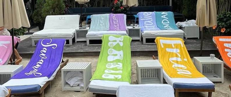 Pu&ograve; includere: Una fila di asciugamani da spiaggia colorati con diverse parole stampate su di essi, tra cui "Summer", "Basking", "Friday", "Italian" e "Brendan". Gli asciugamani sono disposti su sedie a sdraio su una spiaggia di sabbia.
