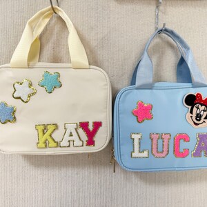 K&ouml;nnte beinhalten: Zwei kleine, rechteckige Handtaschen. Eine ist cremefarben mit sternf&ouml;rmigen Aufn&auml;hern und dem Namen "KAY" in bunten Buchstaben. Die andere ist hellblau und zeigt einen Minnie-Mouse-Aufn&auml;her, einen Stern und den Namen "LUCA". Beide haben Griffe.