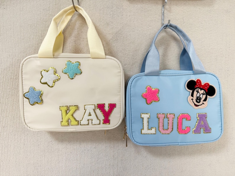 K&ouml;nnte beinhalten: Zwei kleine, rechteckige Handtaschen. Eine ist cremefarben mit sternf&ouml;rmigen Aufn&auml;hern und dem Namen "KAY" in bunten Buchstaben. Die andere ist hellblau und zeigt einen Minnie-Mouse-Aufn&auml;her, einen Stern und den Namen "LUCA". Beide haben Griffe.