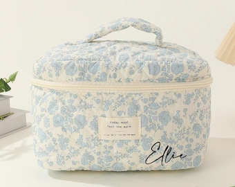 May include: A white quilted makeup bag with a blue floral pattern. The bag has a zipper closure and a top handle. The bag has a small label that says "today, mood feel the warm." and the name "Ellie" is written on the bottom right corner.