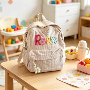 以下が含まれることがあります： ベージュのコーデュロイ製バックパック。フロントポケットにはカラフルな毛糸の文字で「Reese」と刺繍されています。バッグにはトップハンドル、調節可能なショルダーストラップ、フロントジッパーの引き手には白いデイジーのチャームが付いています。