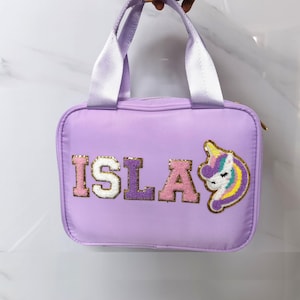 K&ouml;nnte beinhalten: Eine lavendelfarbene Handtasche mit wei&szlig;en Henkeln und einem goldenen Rei&szlig;verschluss. Das Wort "ISLA" ist im College-Stil auf der Vorderseite gestickt, zusammen mit einem Einhorn-Aufn&auml;her mit Regenbogenm&auml;hne und Horn.