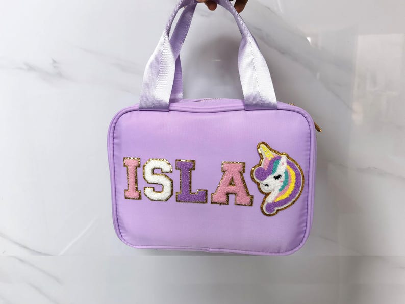 K&ouml;nnte beinhalten: Eine lavendelfarbene Handtasche mit wei&szlig;en Henkeln und einem goldenen Rei&szlig;verschluss. Das Wort "ISLA" ist im College-Stil auf der Vorderseite gestickt, zusammen mit einem Einhorn-Aufn&auml;her mit Regenbogenm&auml;hne und Horn.
