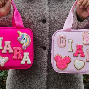 K&ouml;nnte beinhalten: Zwei rechteckige Handtaschen in Pink und Hellrosa mit Griffen. Die pinkfarbene Tasche tr&auml;gt den Namen "CHIARA" mit einem Einhorn- und Blumenaufn&auml;her. Die hellrosa Tasche tr&auml;gt den Namen "GIADA" mit Schleifen-, Blumen-, Herz- und B&auml;renaufn&auml;hern.