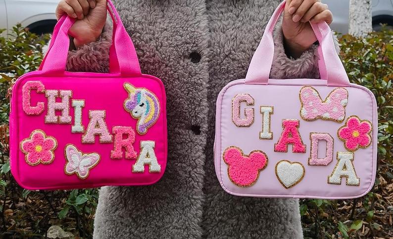 K&ouml;nnte beinhalten: Zwei rechteckige Handtaschen in Pink und Hellrosa mit Griffen. Die pinkfarbene Tasche tr&auml;gt den Namen "CHIARA" mit einem Einhorn- und Blumenaufn&auml;her. Die hellrosa Tasche tr&auml;gt den Namen "GIADA" mit Schleifen-, Blumen-, Herz- und B&auml;renaufn&auml;hern.