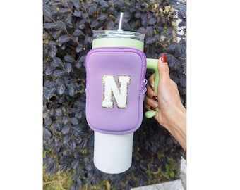 Puede incluir: Portavasos aislante morado con cierre de cremallera y una letra "N" blanca con purpurina dorada. El portavasos está unido a una taza de viaje verde y blanca con asa.