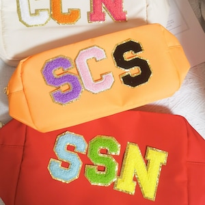 May include: Three colorful zippered pouches with different letter combinations. The pouches are made of a soft, textured fabric and feature large, colorful letters with gold glitter accents. The letters spell out "CCN", "SCS", and "SSN".