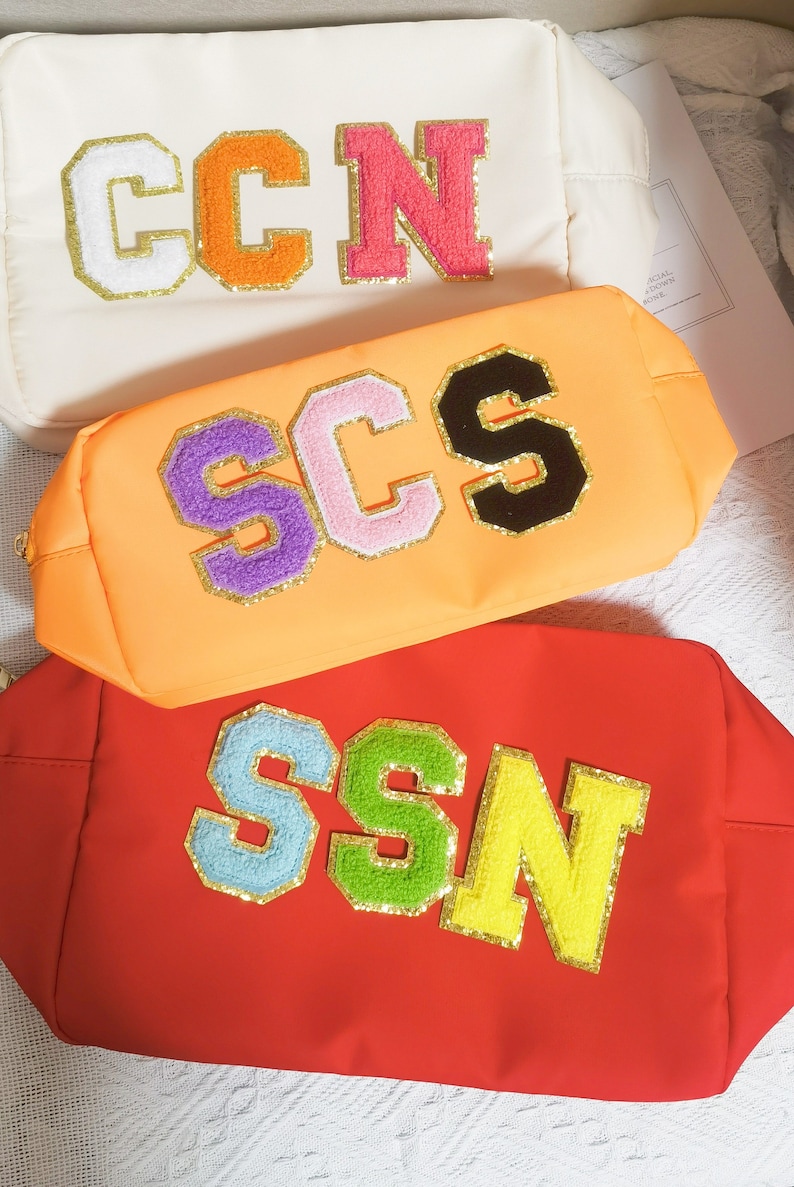 May include: Three colorful zippered pouches with different letter combinations. The pouches are made of a soft, textured fabric and feature large, colorful letters with gold glitter accents. The letters spell out "CCN", "SCS", and "SSN".