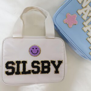 K&ouml;nnte beinhalten: Wei&szlig;e Tragetasche mit einem lila Smiley-Aufn&auml;her und dem Namen "SILSBBY" in schwarzen Buchstaben mit goldenem Rand. Eine hellblaue Tasche mit dem Namen "NATALIE" und einem rosa Stern-Aufn&auml;her ist ebenfalls zu sehen.