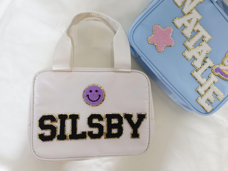 K&ouml;nnte beinhalten: Wei&szlig;e Tragetasche mit einem lila Smiley-Aufn&auml;her und dem Namen "SILSBBY" in schwarzen Buchstaben mit goldenem Rand. Eine hellblaue Tasche mit dem Namen "NATALIE" und einem rosa Stern-Aufn&auml;her ist ebenfalls zu sehen.