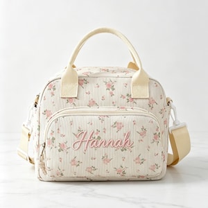 Puede incluir: Un bolso de pana color crema con un delicado estampado floral en rosa y verde. El bolsillo frontal muestra el nombre Hannah bordado en letra cursiva rosa. Incluye dos asas superiores y una correa de hombro color crema ajustable y extraíble.