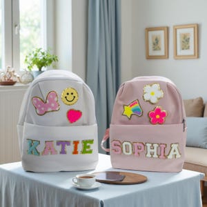Può includere: Due zaini personalizzati, uno bianco e uno rosa, sono su un tavolo. Lo zaino bianco ha il nome "KATIE" e toppe decorative. Lo zaino rosa ha il nome "SOPHIA" e toppe a forma di fiori e stelle. Entrambi hanno tasche frontali.