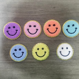 Op de afbeelding: Zeven geborduurde smiley-patches in verschillende kleuren, waaronder paars, roze, oranje, blauw, geel en wit. Elke patch heeft een gouden rand en is ongeveer 5 cm in diameter.