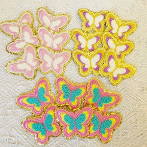 May include: A collection of colorful butterfly patches. The patches feature pink, yellow, and teal wings with gold glitter edging. Some butterflies have pink and white wings, while others have purple and yellow wings.