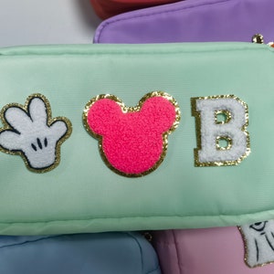 May include: A light green zippered pouch with a white Mickey Mouse hand, a pink Mickey Mouse head, and a white letter "B" with gold glitter trim.