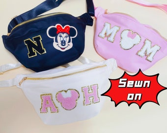 May include: Three fanny packs in navy blue, light pink, and white, each featuring gold zippers and decorative patches. The navy bag has a letter N and Minnie Mouse, the pink bag has MOM with a Minnie icon, and the white bag has A, a Minnie icon, and H.