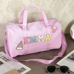 May include: A light pink duffel bag with the name "EMMA" spelled out in colorful, sequined letters. The bag has a shoulder strap and two handles. Decorative patches include a castle and a mouse head. Sunglasses and newspapers are in the foreground.