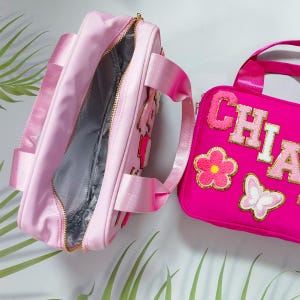 K&ouml;nnte beinhalten: Zwei rosa Lunchpakete. Eines ist offen und zeigt ein graues Innenfutter und einen goldenen Rei&szlig;verschluss. Das andere ist ein helleres Rosa mit dem Namen "CHIA" in goldenen und rosa Buchstaben, zusammen mit Blumen- und Schmetterlingsapplikationen. Beide haben rosa Tr&auml;ger.
