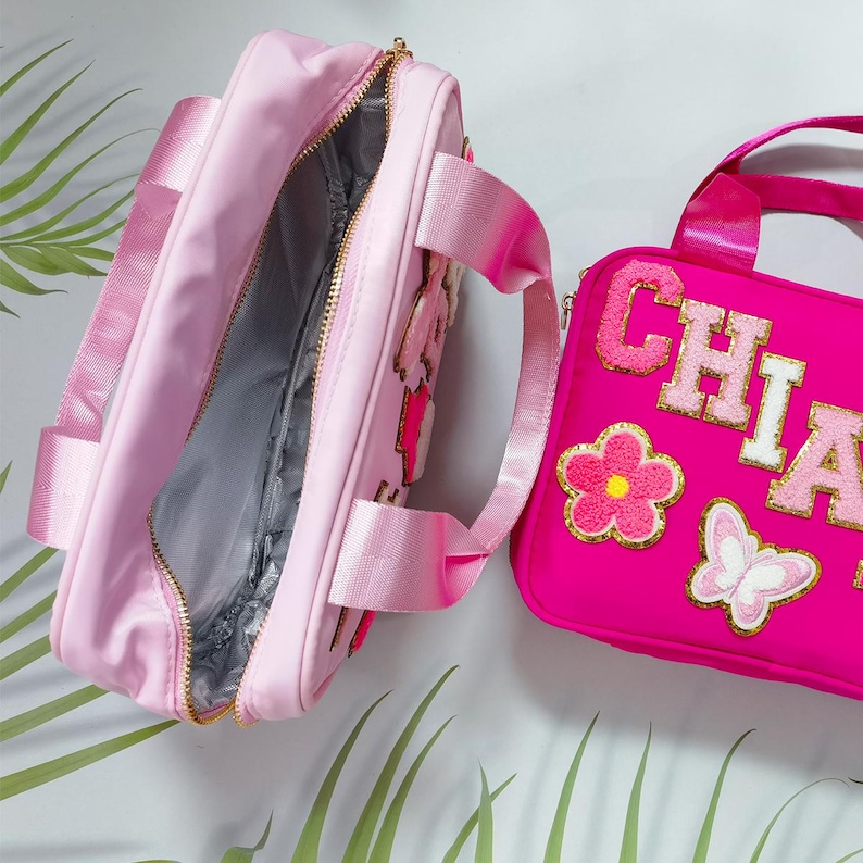 K&ouml;nnte beinhalten: Zwei rosa Lunchpakete. Eines ist offen und zeigt ein graues Innenfutter und einen goldenen Rei&szlig;verschluss. Das andere ist ein helleres Rosa mit dem Namen "CHIA" in goldenen und rosa Buchstaben, zusammen mit Blumen- und Schmetterlingsapplikationen. Beide haben rosa Tr&auml;ger.