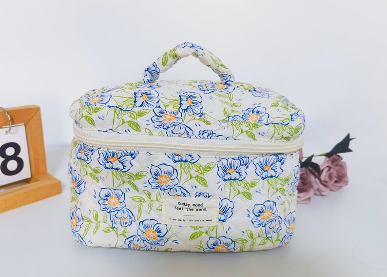 May include: A white cosmetic bag with a floral pattern of blue and orange flowers and green leaves. The bag has a zipper closure and a handle. The text "today, mood feel the warm" is on a small label.
