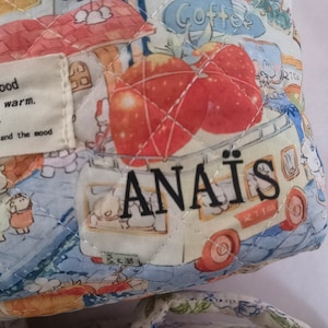 May include: Quilted fabric bag with a whimsical print featuring cartoon characters, apples, and a food truck. The bag has a label that reads "today, mood feel the warm." and the name "ANA&Iuml;S". The bag is in shades of blue, white, red, and yellow.