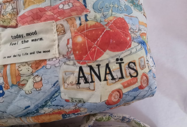May include: Quilted fabric bag with a whimsical print featuring cartoon characters, apples, and a food truck. The bag has a label that reads "today, mood feel the warm." and the name "ANA&Iuml;S". The bag is in shades of blue, white, red, and yellow.