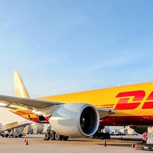 May include: A yellow and red DHL cargo plane with a large engine on the right side. The plane is parked on a tarmac with other cargo planes and trucks in the background.