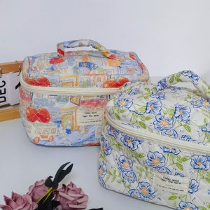 May include: Two quilted cosmetic bags. One features a colorful street scene print, the other a white floral pattern with blue and green accents. Both have a top handle and a zipper closure. The bags have the text "today, mood feel the warm."