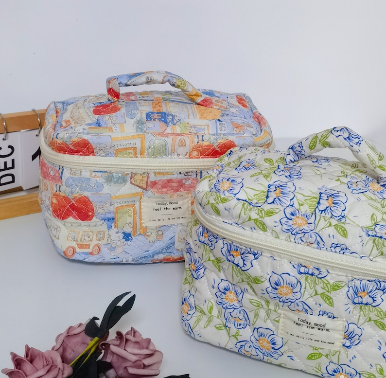 May include: Two quilted cosmetic bags. One features a colorful street scene print, the other a white floral pattern with blue and green accents. Both have a top handle and a zipper closure. The bags have the text "today, mood feel the warm."