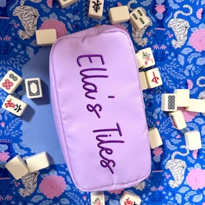May include: A rectangular lavender zippered pouch with the embroidered text "Ella's Tiles" in dark purple thread. The pouch is surrounded by scattered off-white Mahjong tiles featuring various colourful symbols, characters, and floral designs.