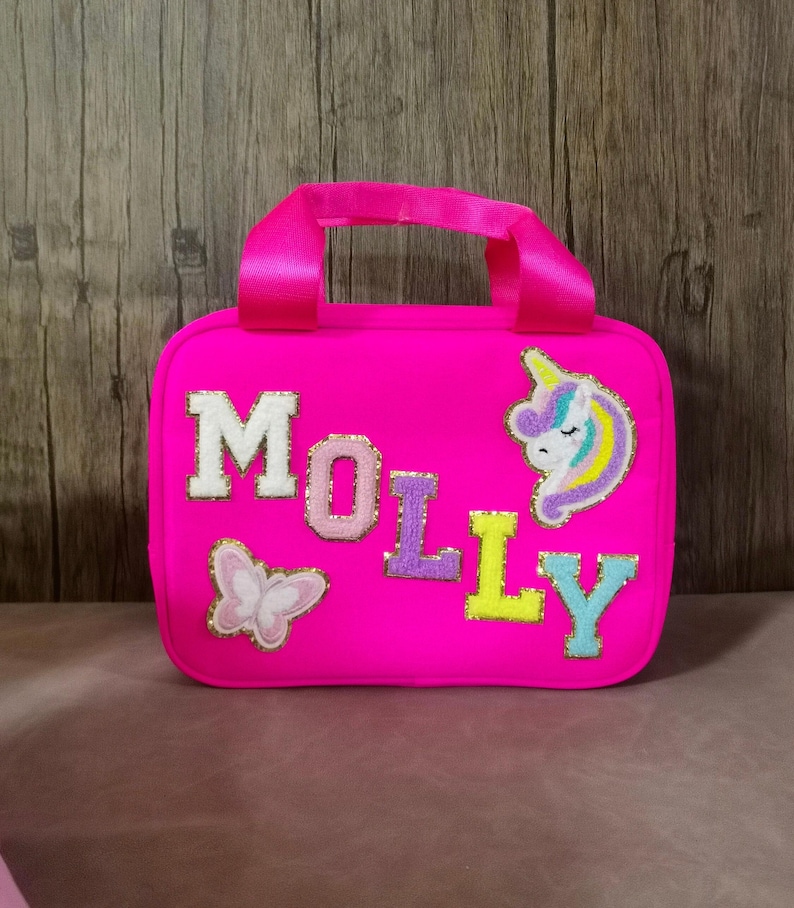 K&ouml;nnte beinhalten: Eine leuchtend pinkfarbene Tragetasche mit passendem Griff. Der Name "MOLLY" ist in bunten, strukturierten Buchstaben auf der Vorderseite geschrieben, zusammen mit einem Schmetterling und einem Einhorn-Aufn&auml;her. Die Tasche hat eine rechteckige Form.