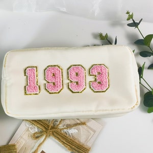 Könnte beinhalten: Weiße Reißverschlusstasche mit den gestickten Zahlen "1993" in Rosa und Gold auf der Vorderseite.