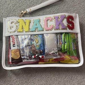 May include: A white, clear pouch filled with snacks, including chocolate bars and sweets. The word "SNACKS" is displayed in colourful, textured letters. The pouch has a wrist strap and gold-coloured hardware.