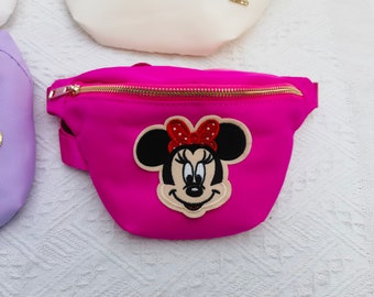 May include: A fuchsia fanny pack with a gold zipper and a Minnie Mouse patch. The patch features Minnie's face with a red bow. Other fanny packs in white and purple are visible in the background.