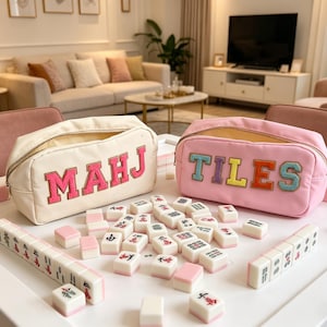 Può includere: Un set da Mahjong bianco e rosa con due astucci con cerniera. Uno è color crema con "MAHJ" a lettere rosa, l'altro è rosa con "TILES" a lettere colorate. Il set è su un tavolo bianco.