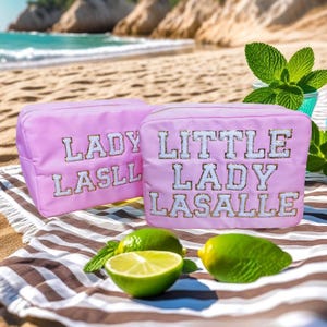 Könnte beinhalten: Zwei rosa Reißverschlusstaschen mit dem Text "Lady LaSalle" und "Little Lady LaSalle" in weißen Buchstaben mit goldenen Glitzerakzenten. Die Taschen liegen auf einem gestreiften Strandtuch mit Limetten.