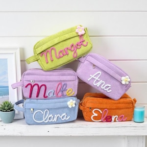 Puede incluir: Bolsas de cosméticos de pana personalizadas en varios colores, como verde, morado, azul y naranja. Cada bolsa tiene un nombre bordado en un color contrastante y una flor decorativa.