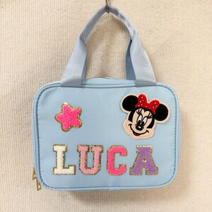 K&ouml;nnte beinhalten: Hellblaue Tragetasche mit Henkel, verziert mit einem Minnie Mouse-Aufn&auml;her und dem Namen "LUCA" in bunten, glitzernden Buchstaben. Ein pinkfarbener sternf&ouml;rmiger Aufn&auml;her ist ebenfalls auf der Tasche. Die Tasche h&auml;ngt an einem Metallhaken.