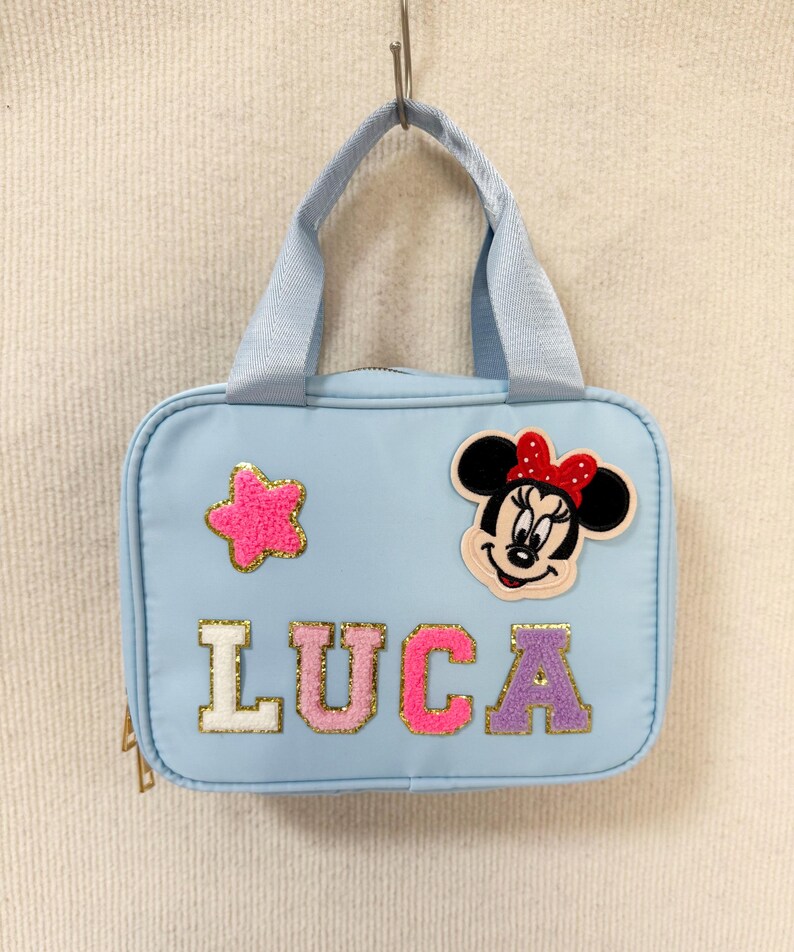 K&ouml;nnte beinhalten: Hellblaue Tragetasche mit Henkel, verziert mit einem Minnie Mouse-Aufn&auml;her und dem Namen "LUCA" in bunten, glitzernden Buchstaben. Ein pinkfarbener sternf&ouml;rmiger Aufn&auml;her ist ebenfalls auf der Tasche. Die Tasche h&auml;ngt an einem Metallhaken.