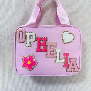 K&ouml;nnte beinhalten: Hellrosa Tragetasche mit passendem Griff. Die Tasche tr&auml;gt den Namen "OPHELIA" in rosa und wei&szlig;en Buchstaben, sowie eine Blume und ein Herz. Die Applikationen sind goldfarben umrandet.