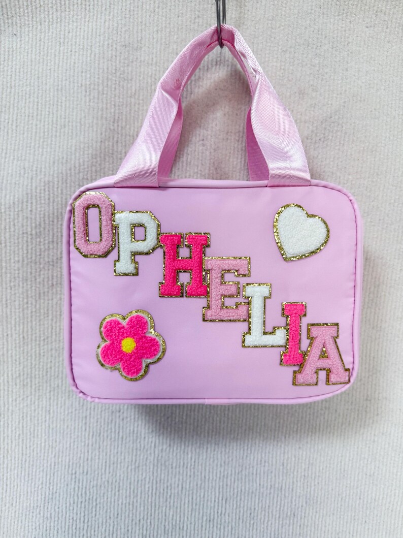 K&ouml;nnte beinhalten: Hellrosa Tragetasche mit passendem Griff. Die Tasche tr&auml;gt den Namen "OPHELIA" in rosa und wei&szlig;en Buchstaben, sowie eine Blume und ein Herz. Die Applikationen sind goldfarben umrandet.