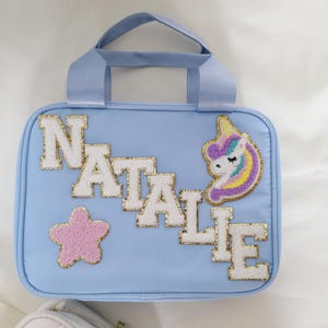 K&ouml;nnte beinhalten: Hellblaue Handtasche mit oberem Griff, mit dem Namen "NATALIE" in wei&szlig;en und goldglitzernden Buchstaben. Ein rosa Stern und ein Einhorn-Aufn&auml;her verzieren die Tasche. Die Tasche ist rechteckig mit abgerundeten Ecken.