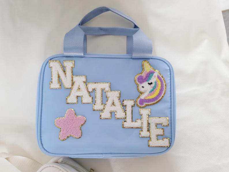 K&ouml;nnte beinhalten: Hellblaue Handtasche mit oberem Griff, mit dem Namen "NATALIE" in wei&szlig;en und goldglitzernden Buchstaben. Ein rosa Stern und ein Einhorn-Aufn&auml;her verzieren die Tasche. Die Tasche ist rechteckig mit abgerundeten Ecken.