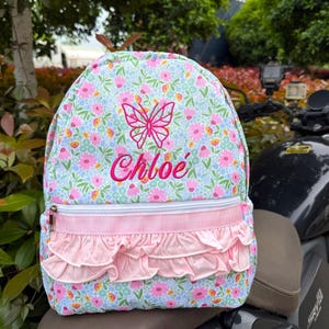 Puede incluir: Una mochila con estampado floral con una mariposa rosa y el nombre "Chloe" bordado en rosa. La mochila presenta un ribete con volantes rosas y una cremallera blanca.