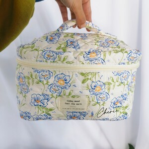 May include: A quilted cosmetic bag with a floral pattern in blue and white. The bag has a zipper closure and a handle. The text "today, mood feel the warm" and "Cherie" are printed on the bag.