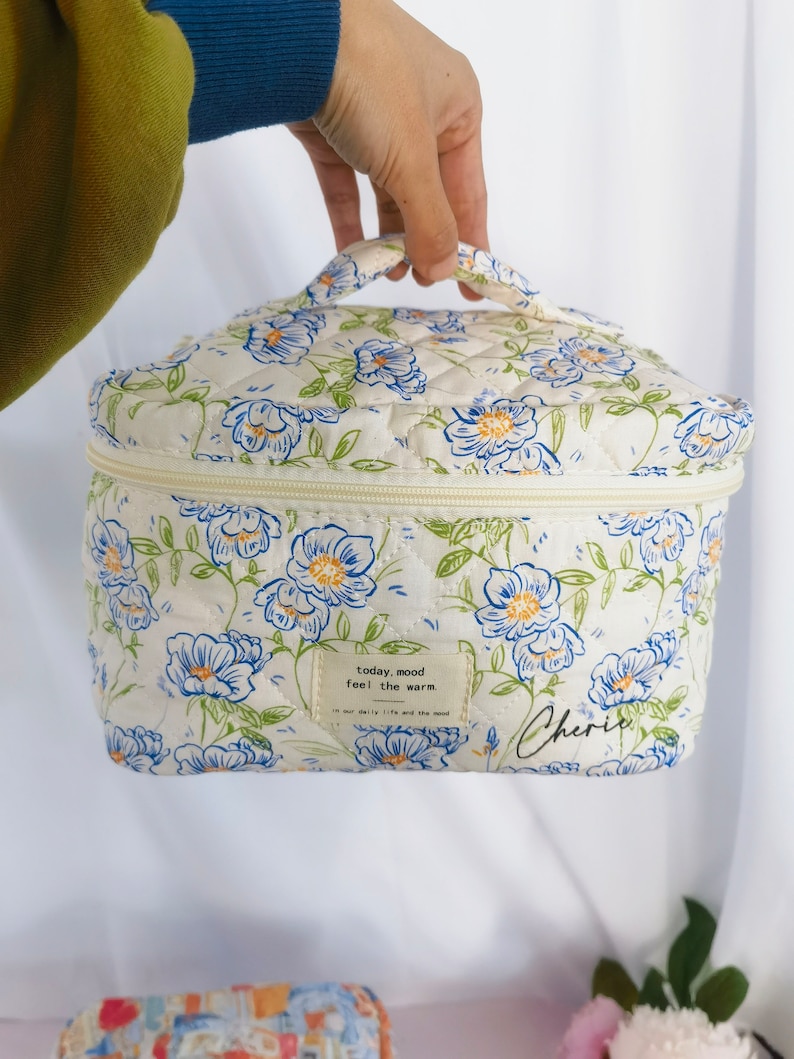May include: A quilted cosmetic bag with a floral pattern in blue and white. The bag has a zipper closure and a handle. The text "today, mood feel the warm" and "Cherie" are printed on the bag.