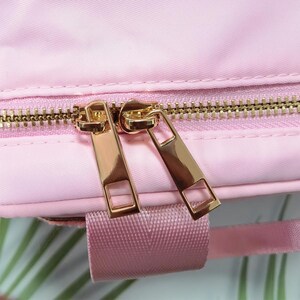 K&ouml;nnte beinhalten: Nahaufnahme eines rosa Stoffes mit einem goldenen Rei&szlig;verschluss. Der Rei&szlig;verschluss hat zwei goldene Zipper. Ein rosa Gurt ist am unteren Bildrand zu sehen. Der Stoff scheint eine Tasche oder ein &auml;hnlicher Artikel zu sein.