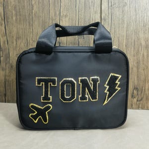 K&ouml;nnte beinhalten: Schwarze Reisetasche mit einem oberen Griff und einem goldenen Rei&szlig;verschluss. Die Tasche zeigt das Wort "TON" in goldenen, umrandeten Buchstaben, zusammen mit einem goldenen Flugzeug und Blitz-Grafik. Die Tasche ist rechteckig und hat einen schwarzen Riemen.